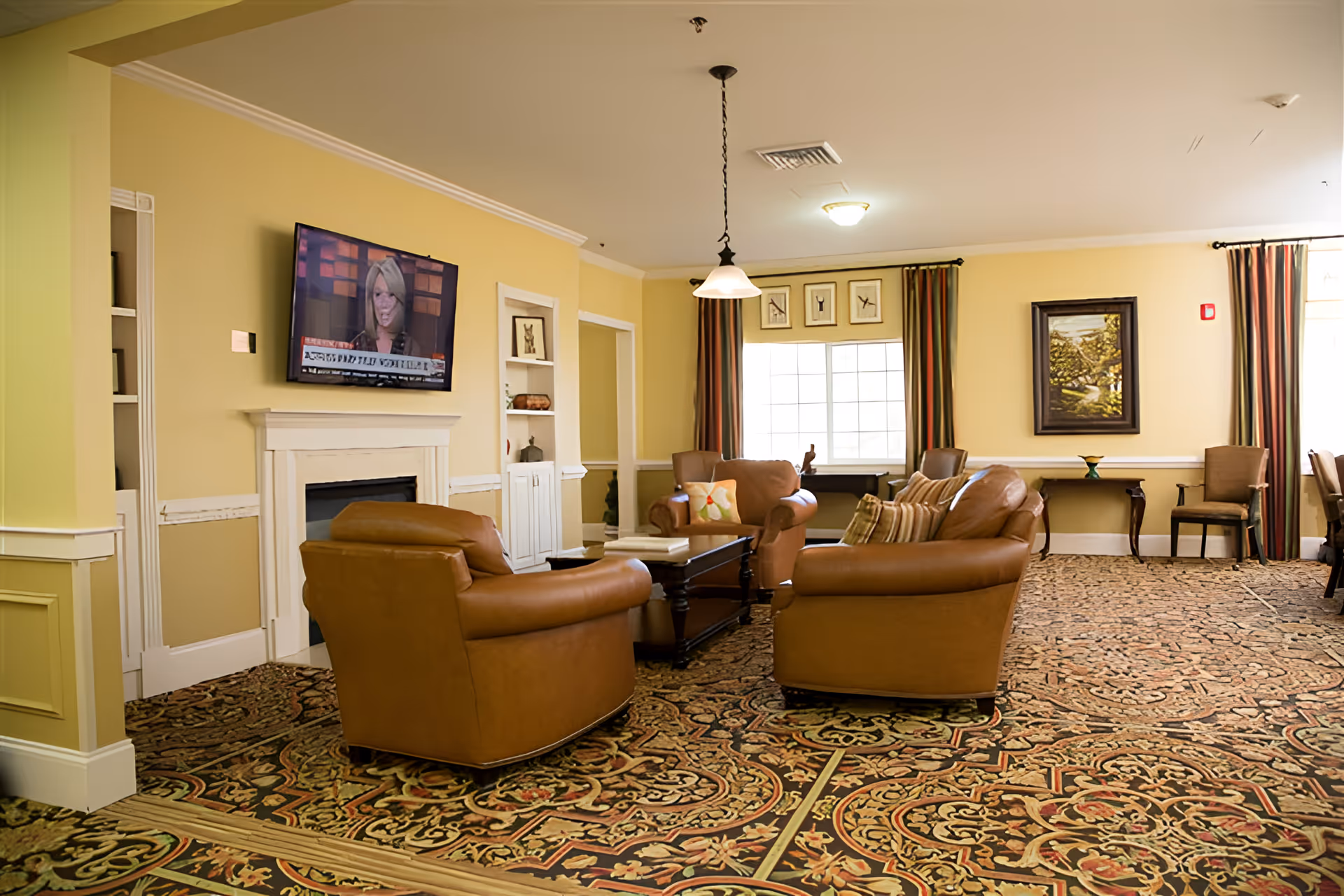 A cozy living room area with three brown leather armchairs arranged around a dark wooden coffee table. The room has a patterned carpet, light yellow walls, a white fireplace with a flat-screen TV mounted above it, and large windows with curtains letting in natural light. There are framed pictures on the walls and additional chairs along the perimeter.