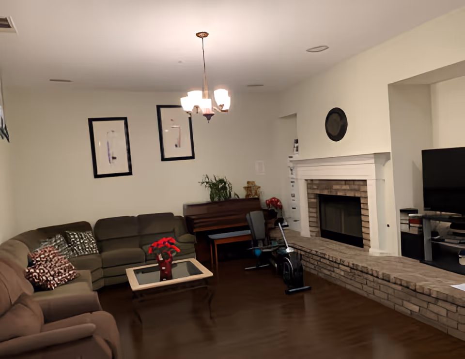 A cozy living room with a sectional sofa, a glass coffee table with a red flower vase, a piano against the wall, an exercise bike, a fireplace with a brick hearth, a flat-screen TV on a stand, and two framed abstract artworks on the wall. The room has wooden flooring and a ceiling light fixture.