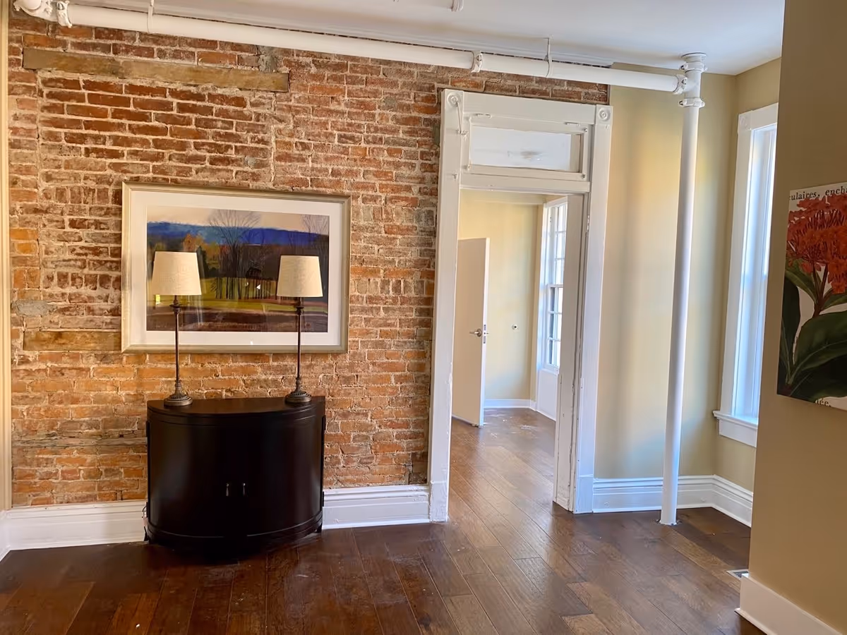 A living room interior with an exposed brick wall, a dark console table topped with two lamps and framed artwork, hardwood floors and a doorway to another room.