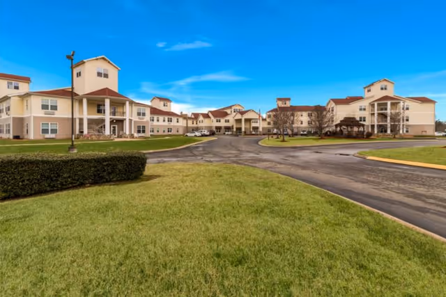 Exterior view of a large senior living facility building with beige walls and red roofs under a clear blue sky. The building surrounds a paved driveway with green grass and trimmed bushes in the foreground.