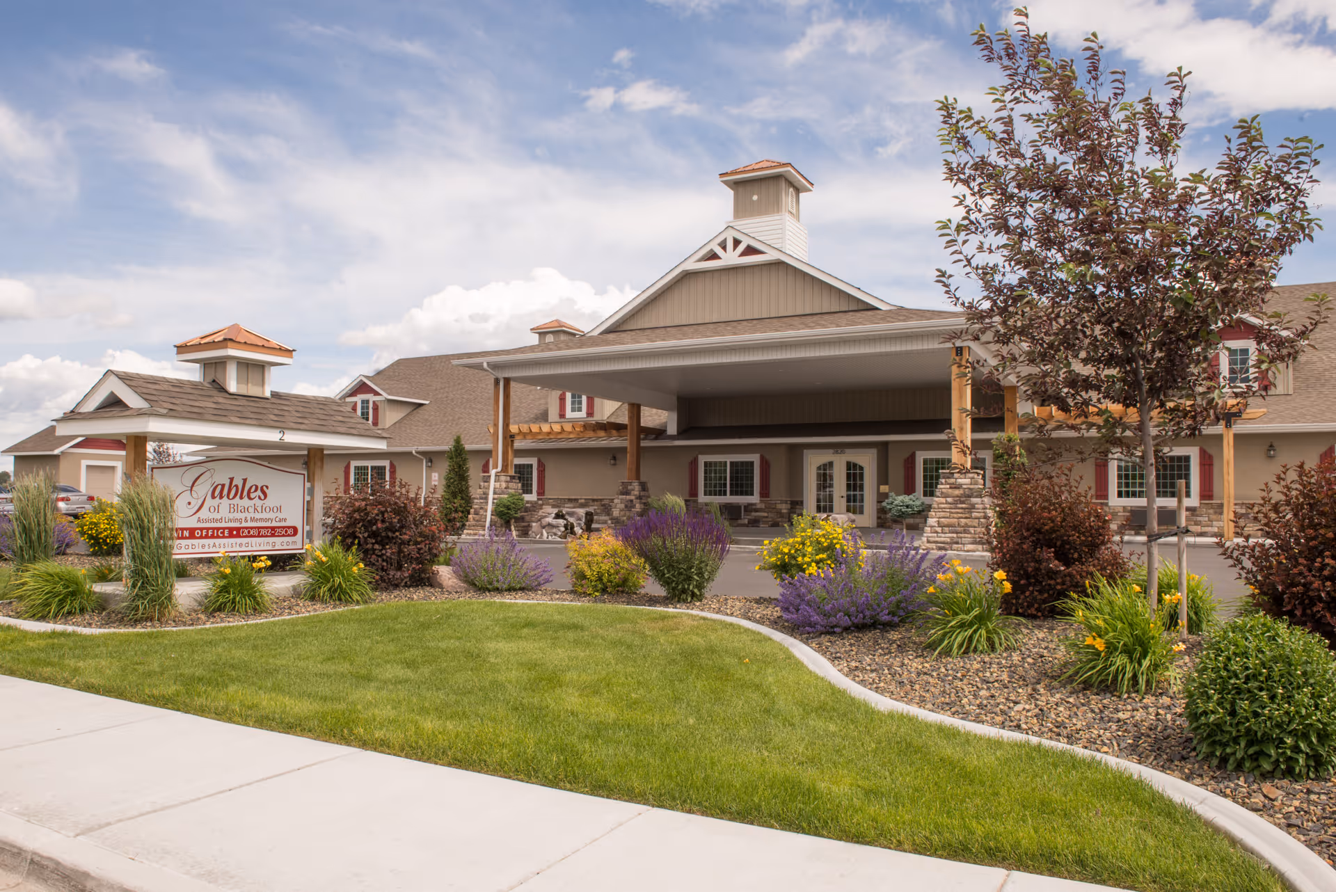 Exterior view of The Gables Assisted Living of Blackfoot building with a covered entrance, landscaped garden with green grass, bushes, and flowers, and a sign displaying the facility's name and contact information.