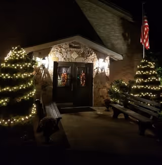 Nighttime view of a stone building entrance decorated with two Christmas wreaths on double doors, flanked by two lit wall lanterns. Two Christmas trees adorned with white string lights are on either side of the entrance, with benches nearby and an American flag on a pole to the right.
