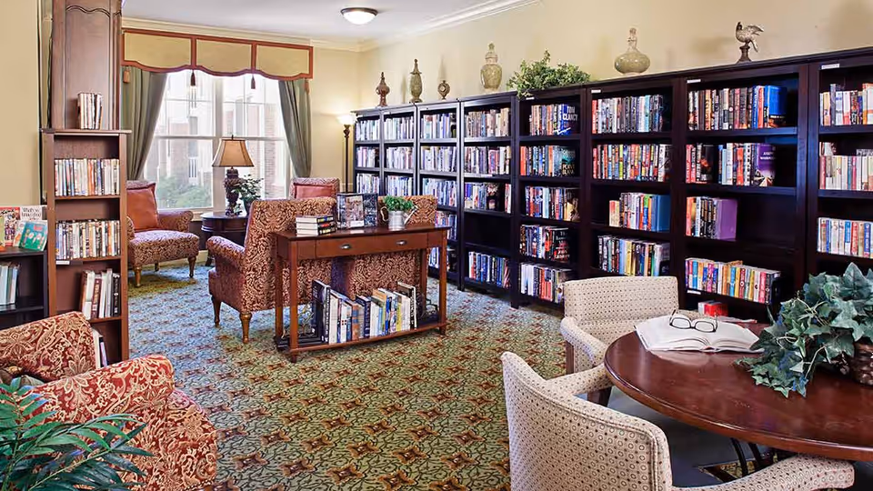 A cozy library room with multiple dark wood bookshelves filled with books, patterned armchairs, a round wooden table with two upholstered chairs, and a window with green curtains letting in natural light. The room has a patterned carpet and decorative items on top of the bookshelves.