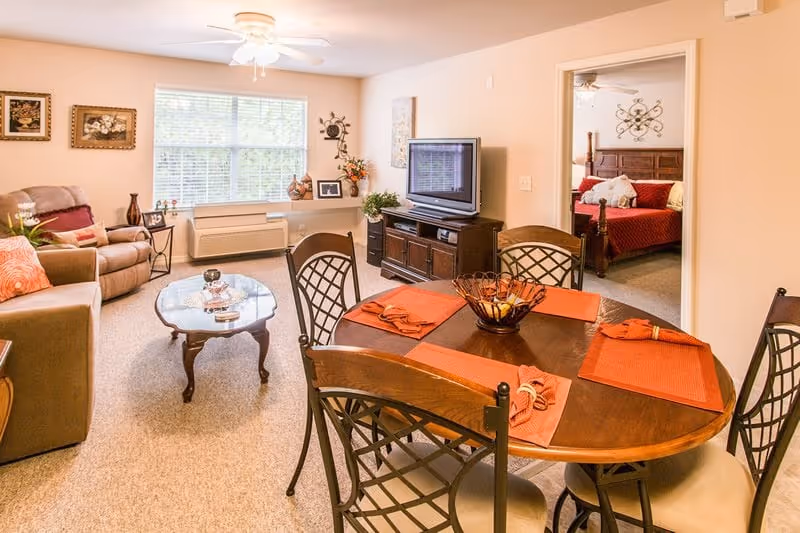 Open-plan living and dining area with a round table set for four, sofa and TV, and a bedroom visible through a doorway.
