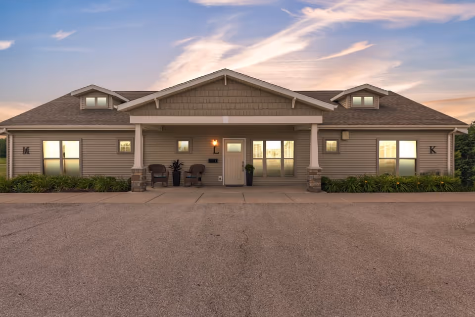 Exterior front view of a single-story residential building with beige siding, a gabled roof, and two small dormer windows. The entrance has a covered porch with two chairs and potted plants. The building is surrounded by a paved area and some greenery along the foundation. The sky is partly cloudy with a warm sunset glow.