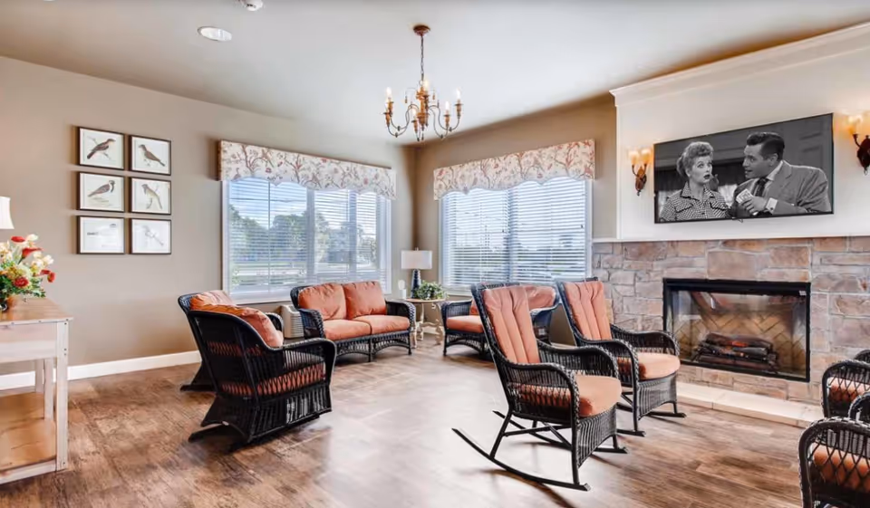 A cozy living room area with wicker furniture featuring orange cushions, a stone fireplace with a mounted TV showing a black and white scene, large windows with floral valances, a chandelier, and framed bird artwork on the wall.