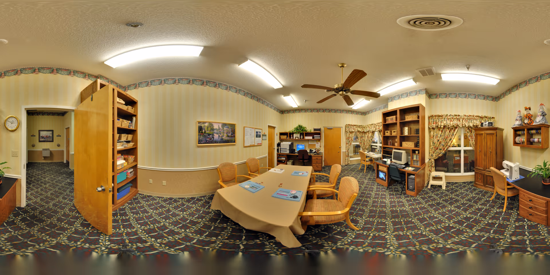 A senior living facility common room with a central table and chairs, bookshelves, computers, and patterned carpet.