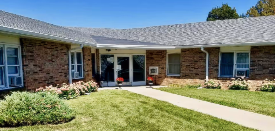 Exterior view of a single-story brick building with a gray shingled roof, a concrete walkway leading to double glass doors, and flower beds with green shrubs and blooming flowers on either side of the entrance.
