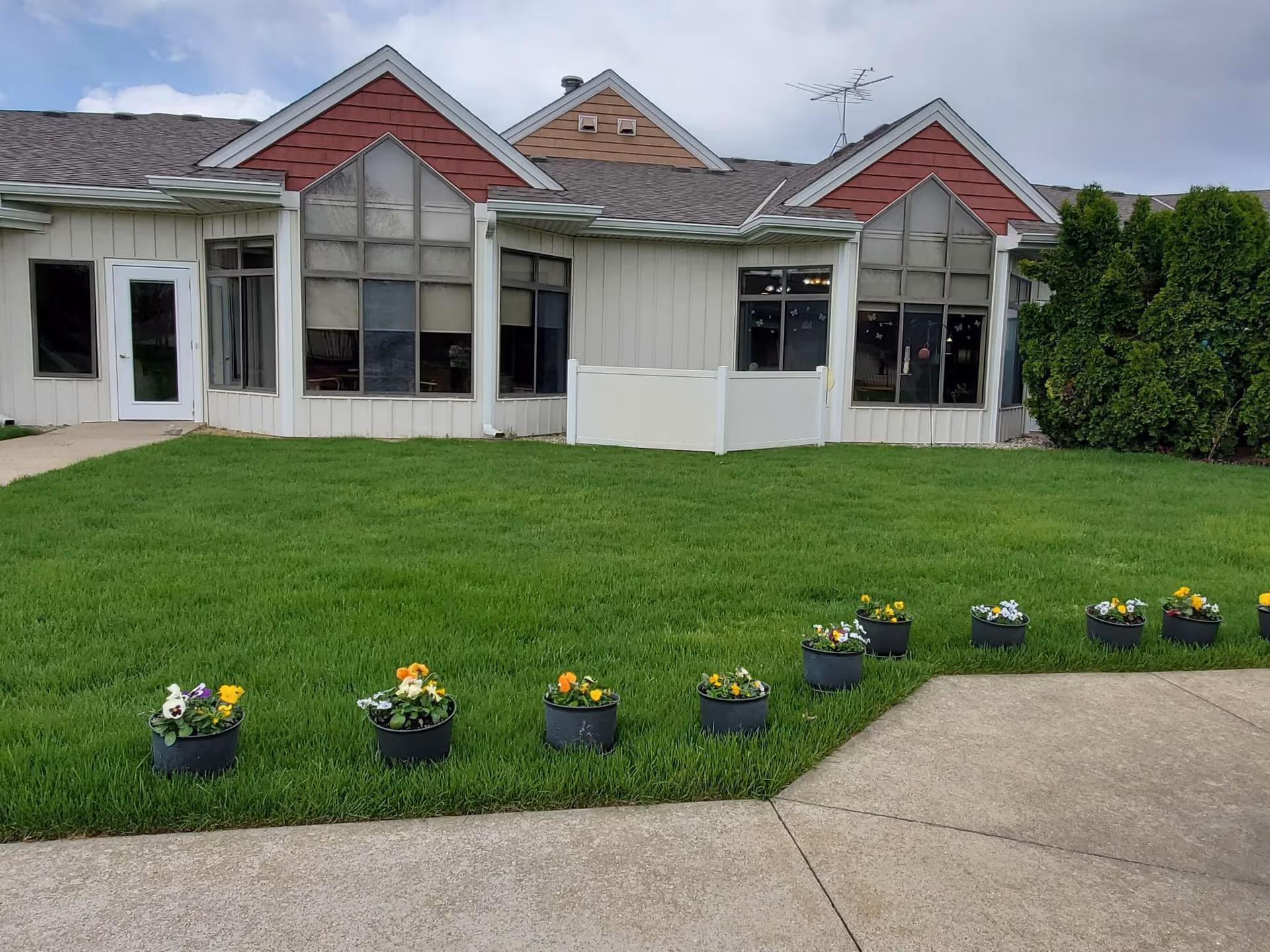 Exterior view of a single-story building with large triangular windows and red and beige siding. In front of the building is a well-maintained green lawn with a row of small flower pots containing colorful flowers. A concrete pathway runs along the lawn.