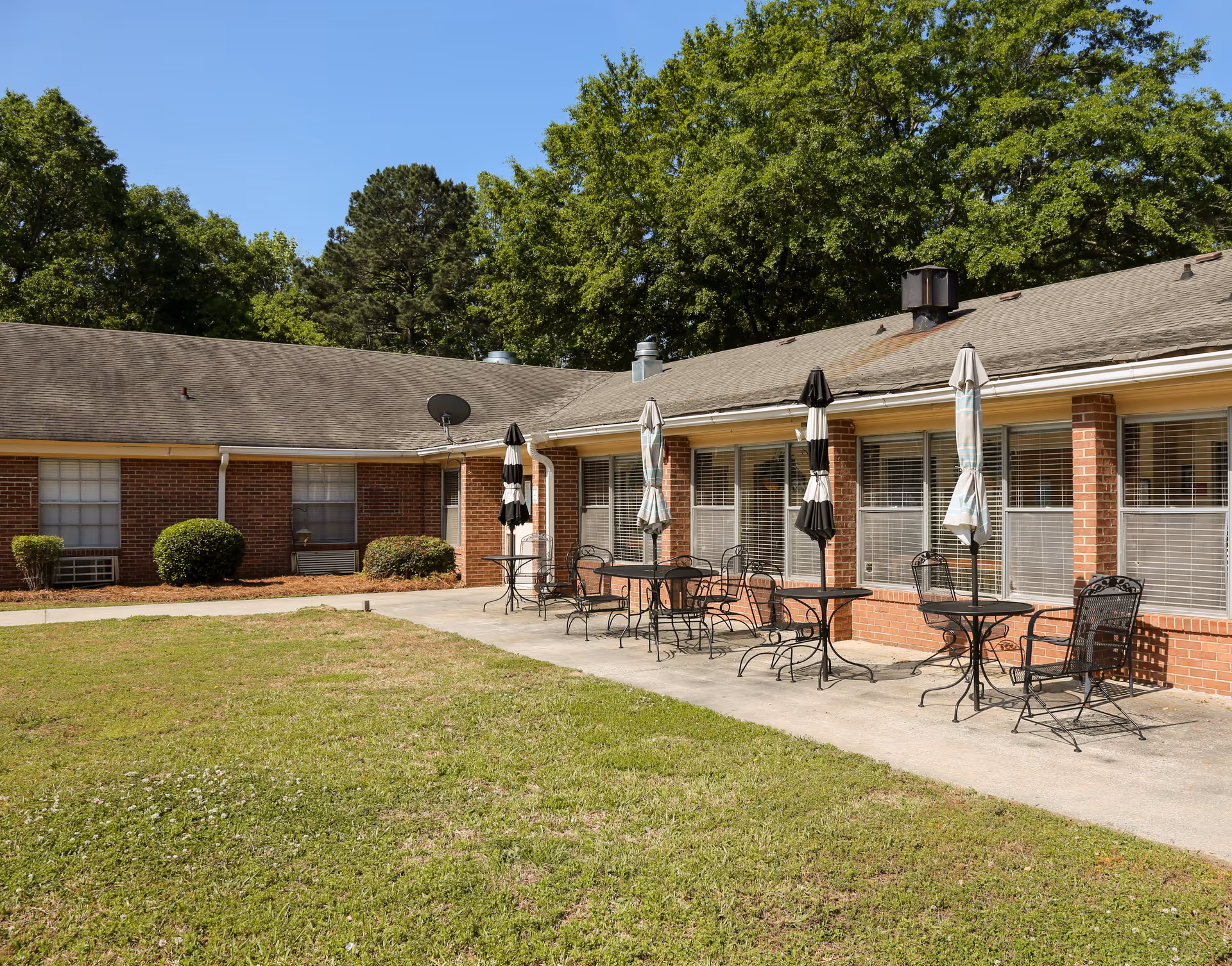 Outdoor patio area at Chaplinwood Health & Rehabilitation with several black metal tables and chairs, each table having a closed umbrella. The patio is adjacent to a brick building with multiple windows and surrounded by green grass and trees under a clear blue sky.
