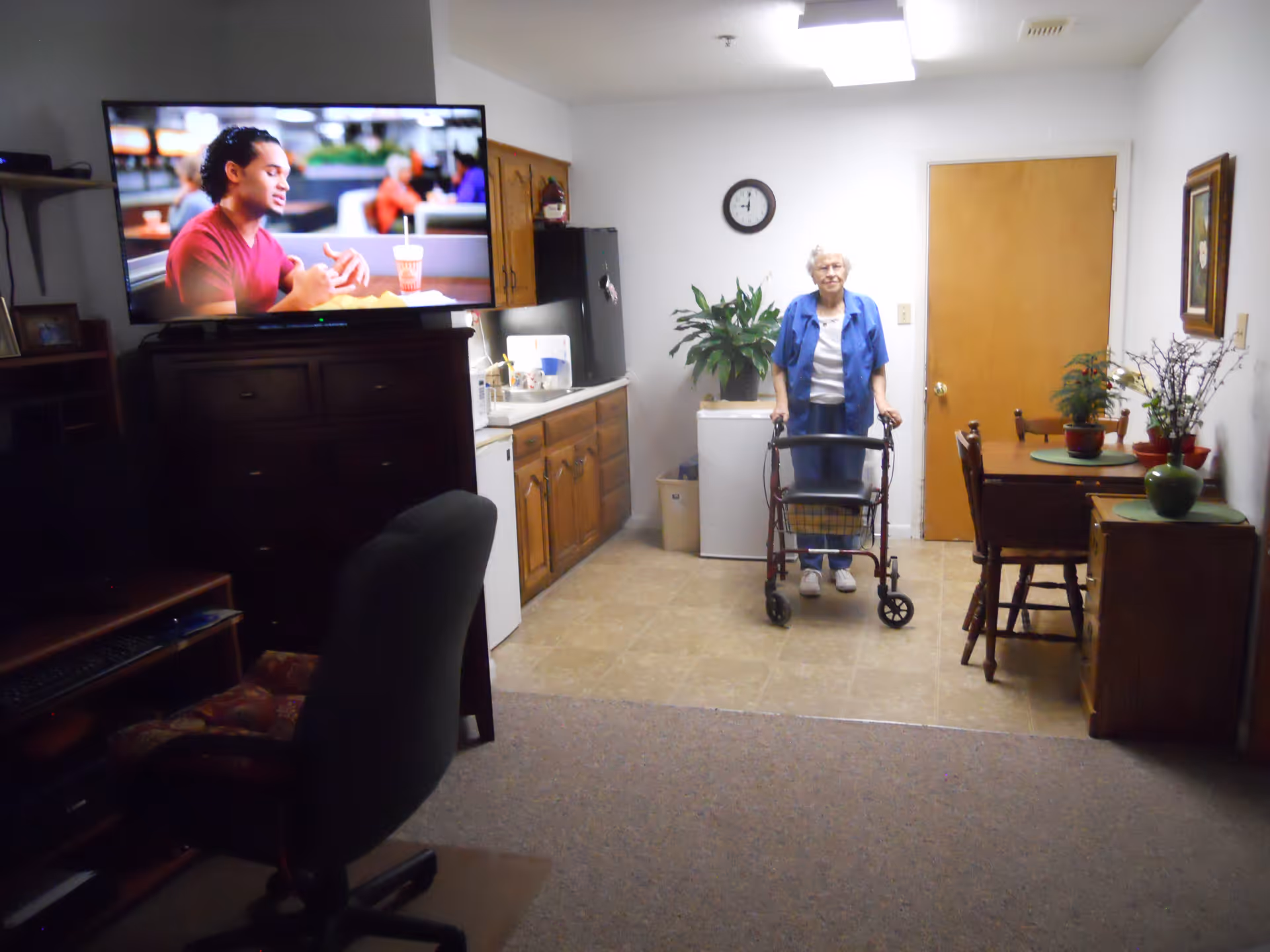 An elderly woman with a walker stands in a room that combines a small kitchen and dining area. The kitchen has wooden cabinets, a refrigerator, and a microwave. A dining table with chairs and potted plants is on the right side. A television mounted on a dark wooden cabinet shows a man speaking. There is a clock on the wall above the woman and a closed wooden door behind her.