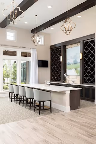 Bright and modern interior space featuring a long white island countertop with six cushioned chairs. The room has large windows with white curtains, dark wood beams on the ceiling, decorative pendant lights, and built-in dark wood wine racks on the wall. A flat-screen TV is mounted on the wall near the windows.