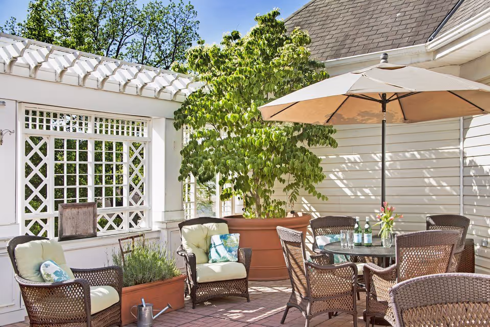 Outdoor patio area with wicker chairs and cushions arranged around a glass-top table under a large beige umbrella. There are potted plants, including a large leafy tree in a terracotta pot, and a white lattice fence with greenery in the background.