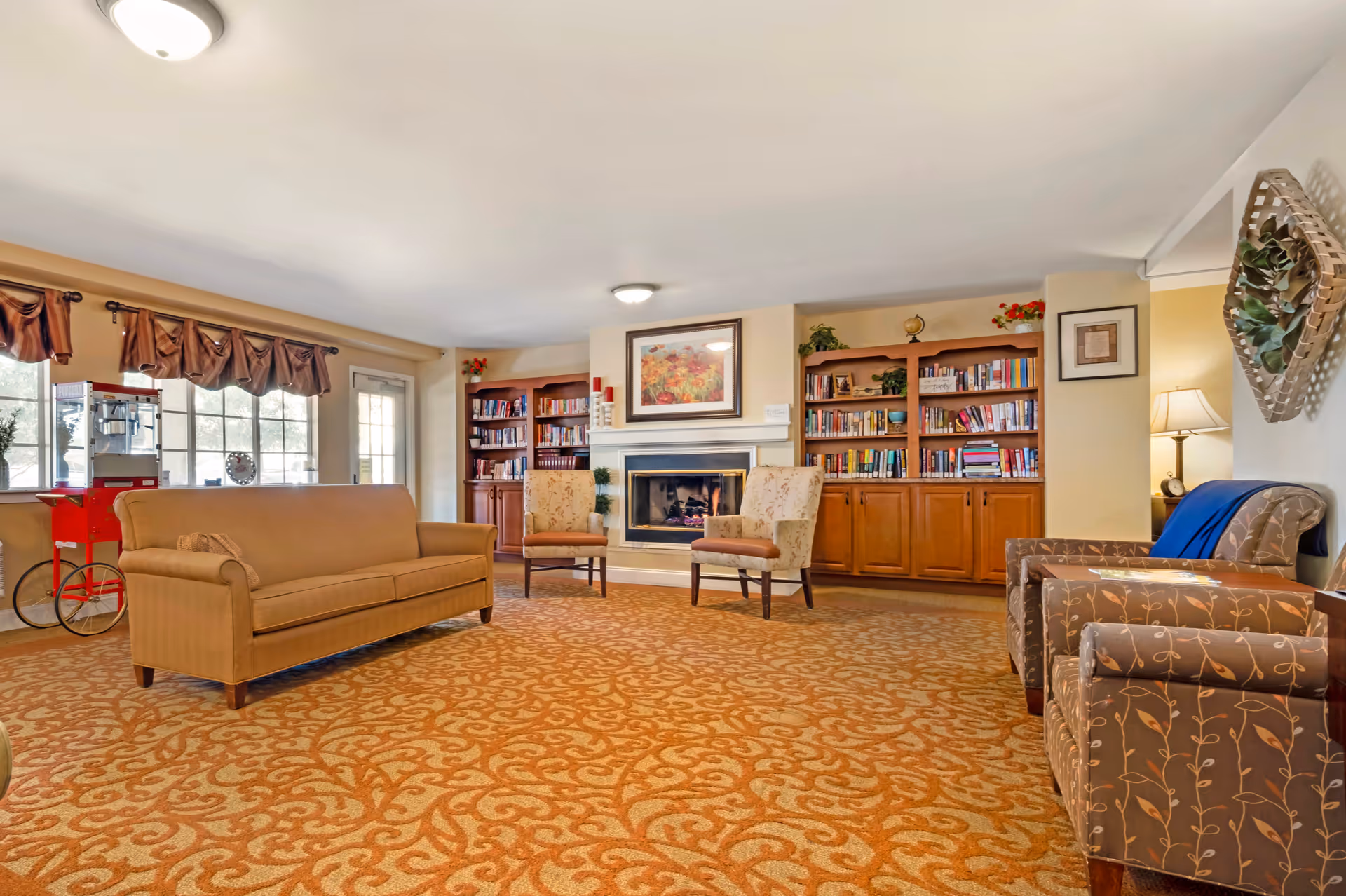 A cozy living room area in a senior living facility featuring a patterned orange carpet, a beige sofa, two floral armchairs, and two brown armchairs with leaf designs. The room has a fireplace with a floral painting above it, flanked by wooden bookshelves filled with books and decorative plants. There is a popcorn machine near the windows with brown valances, and a lamp on a side table next to one of the armchairs. A decorative wall basket with greenery hangs on the right wall.