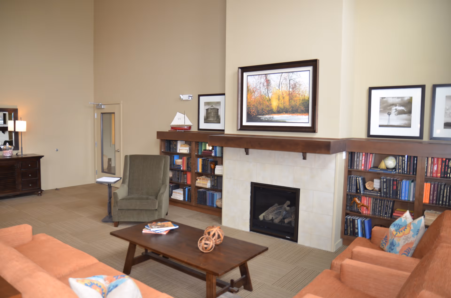 A cozy living room with a fireplace surrounded by built-in bookshelves filled with books and decorative items. There are two orange armchairs with colorful pillows and a green armchair near a small side table. A wooden coffee table with decorative objects and magazines sits in the center. Above the fireplace is a framed picture of a forest scene in autumn colors, and additional framed black and white photos are on the walls. The room has beige walls and carpeted flooring.