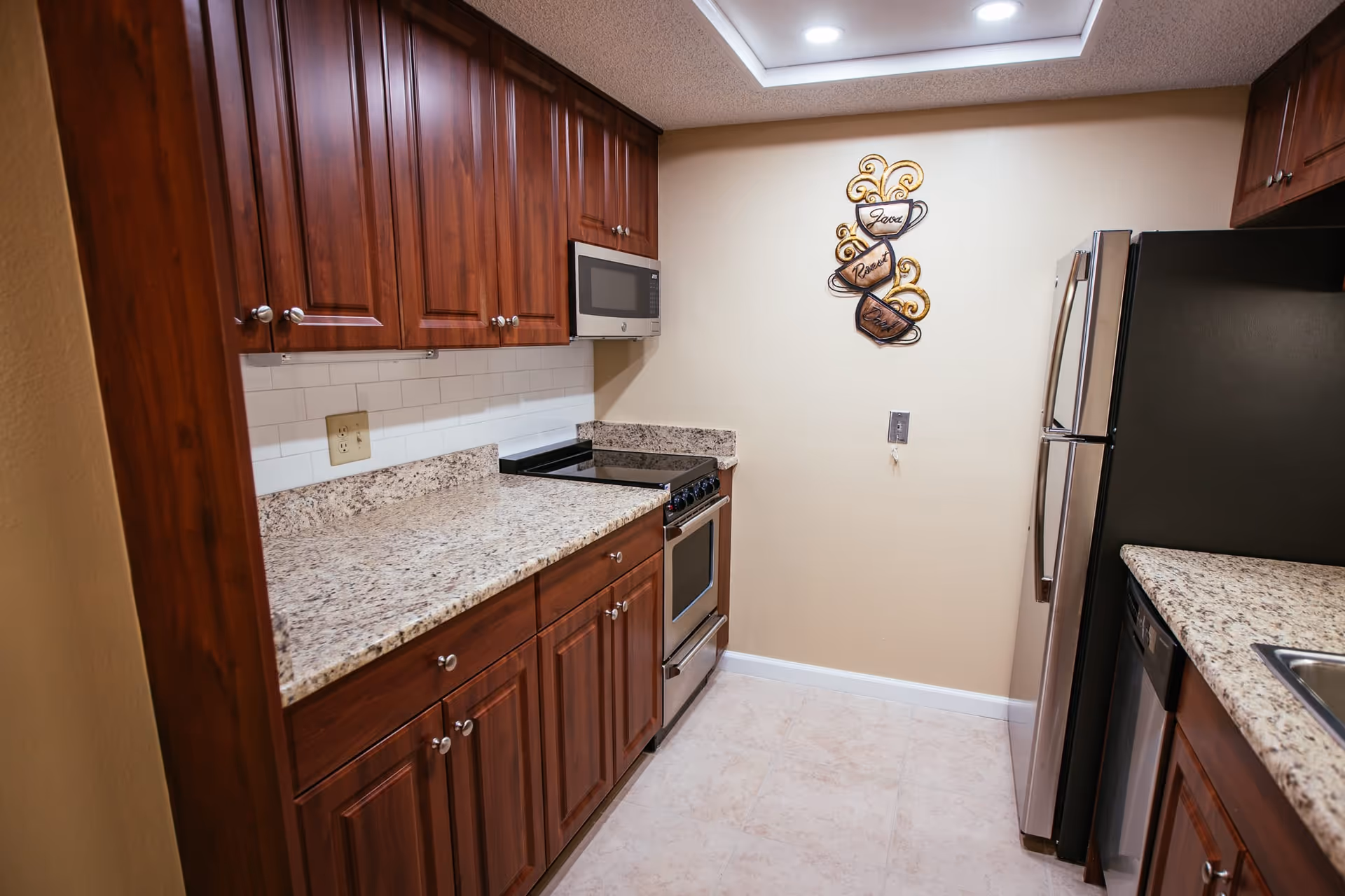 A compact kitchen with wooden cabinets, granite countertops, a stainless steel refrigerator, stove, microwave, and dishwasher. The walls are beige with a decorative metal wall art featuring three coffee cups labeled Java, Roast, and Brew. The floor is tiled and the ceiling has recessed lighting.