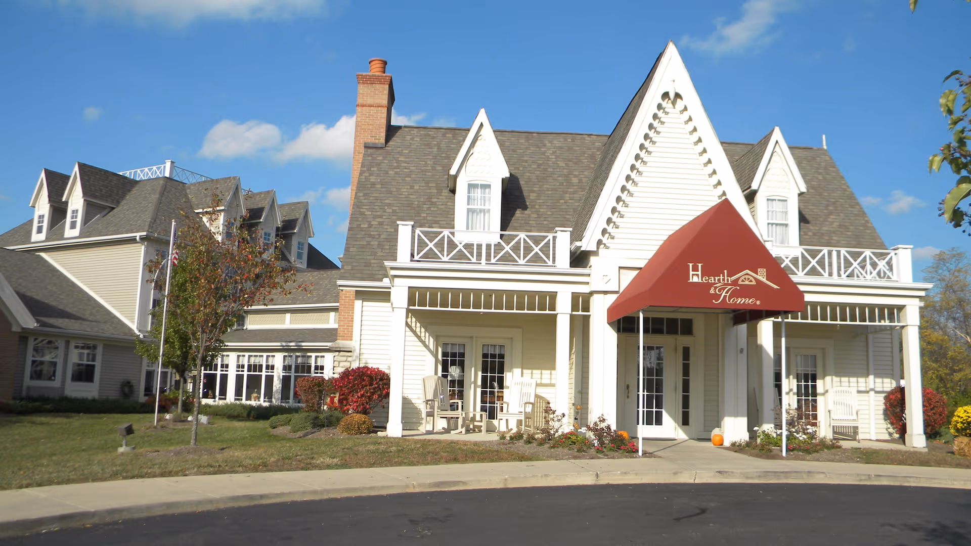 Front entrance of a white two-story senior living building with a red 'Hearth & Home' awning and a porch with rocking chairs.