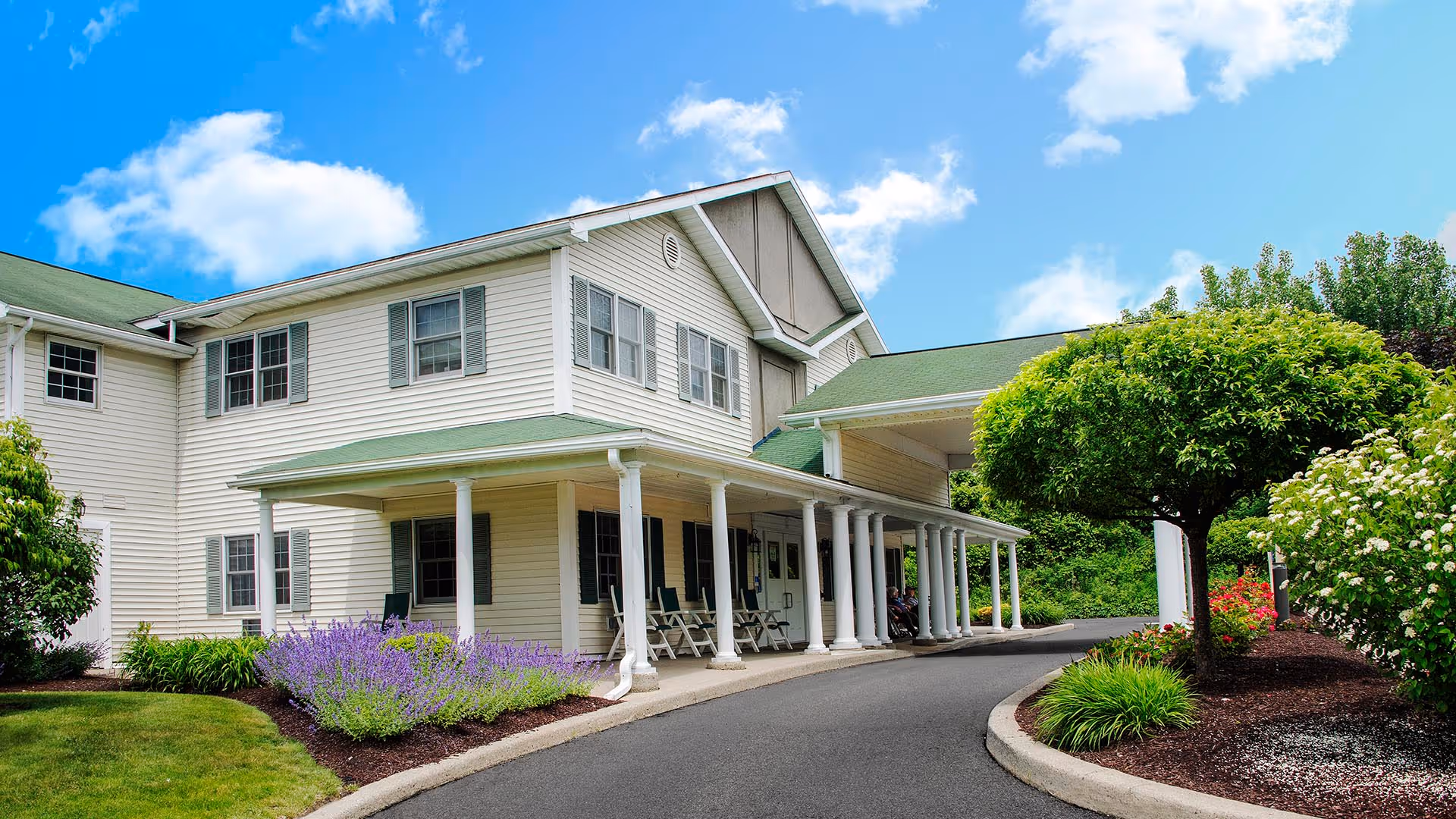 Two-story white senior living building with a covered porch and columns, a curved driveway, and landscaped grounds with flowering shrubs under a blue sky.