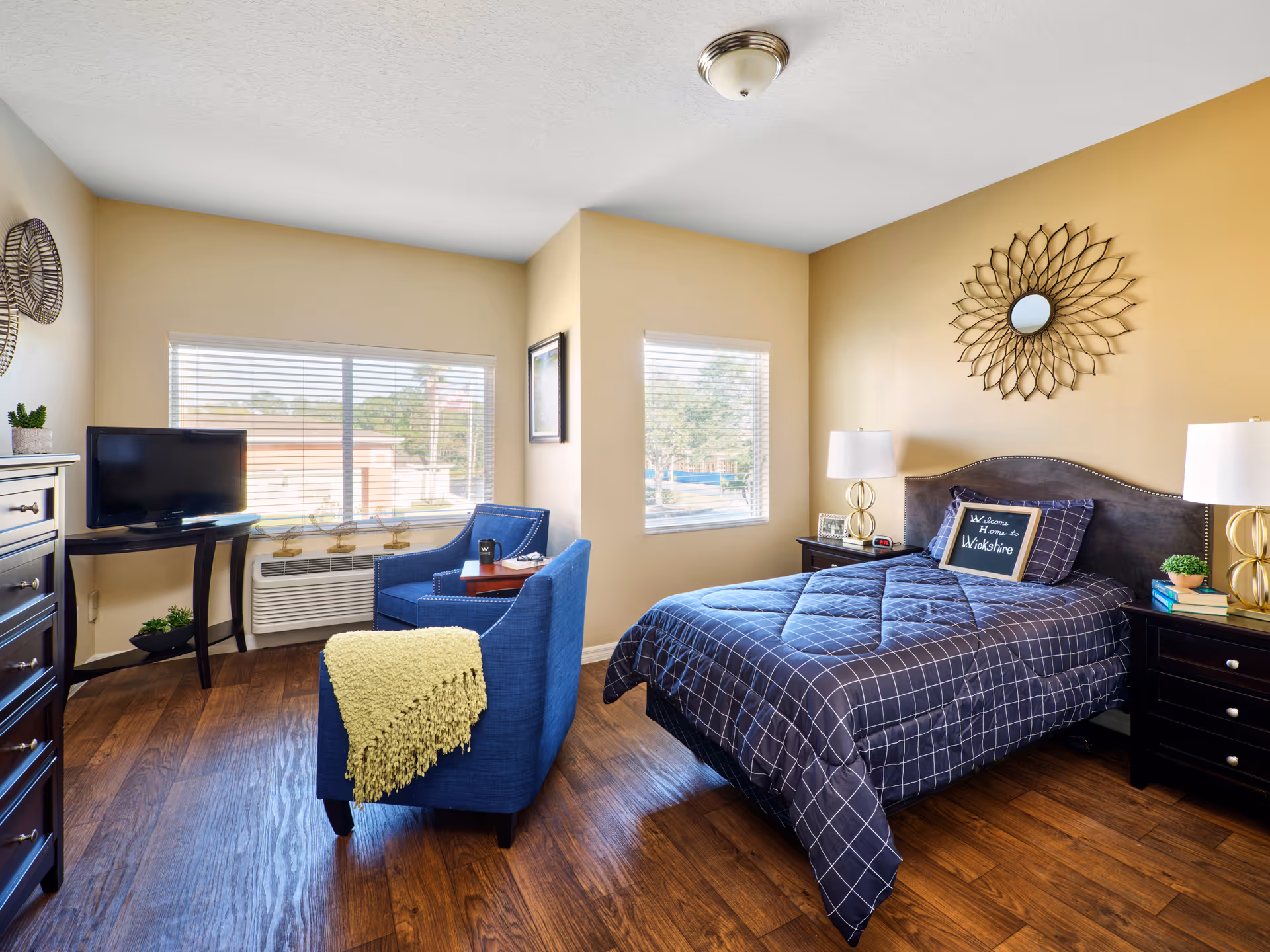 A cozy bedroom with a bed covered in a blue checkered comforter and a pillow that says 'Welcome Home to Wiltshire.' The room has two windows with blinds, a blue armchair with a yellow throw blanket, a small table, a TV on a stand, two nightstands with lamps, and decorative wall art above the bed.