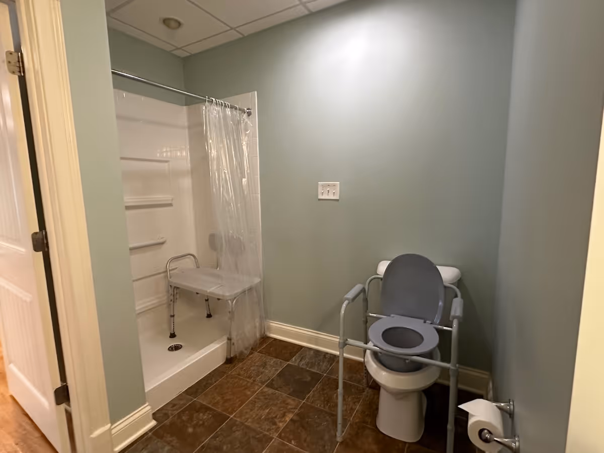 A bathroom with a walk-in shower that has a clear plastic curtain and a white shower chair inside. There is a toilet with a raised seat and arm supports for accessibility. The walls are painted light green and the floor is covered with brown tiles.