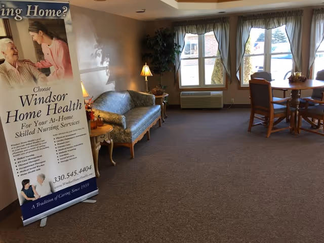 A cozy living room area with a patterned blue sofa, a small side table with a lamp, a large window with curtains letting in natural light, and a round wooden table with chairs. There is a standing banner on the left side advertising Windsor Home Health skilled nursing services.