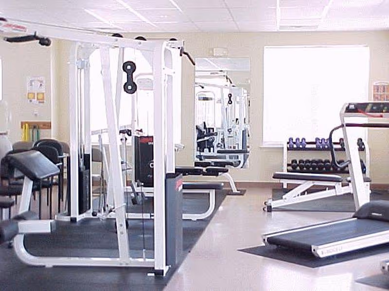 A fitness room with various exercise equipment including a treadmill, weight machines, and free weights. The room has light-colored walls, a large window, and mirrors on the wall.