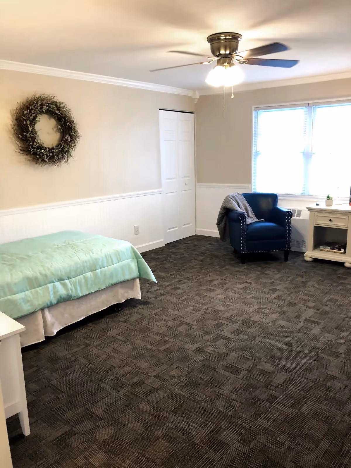 A senior living bedroom with a single bed covered in a light green blanket, a blue armchair with a gray throw draped over it, a white nightstand with a small plant and books, a window with blinds, a ceiling fan with lights, and a decorative wreath on the beige wall.