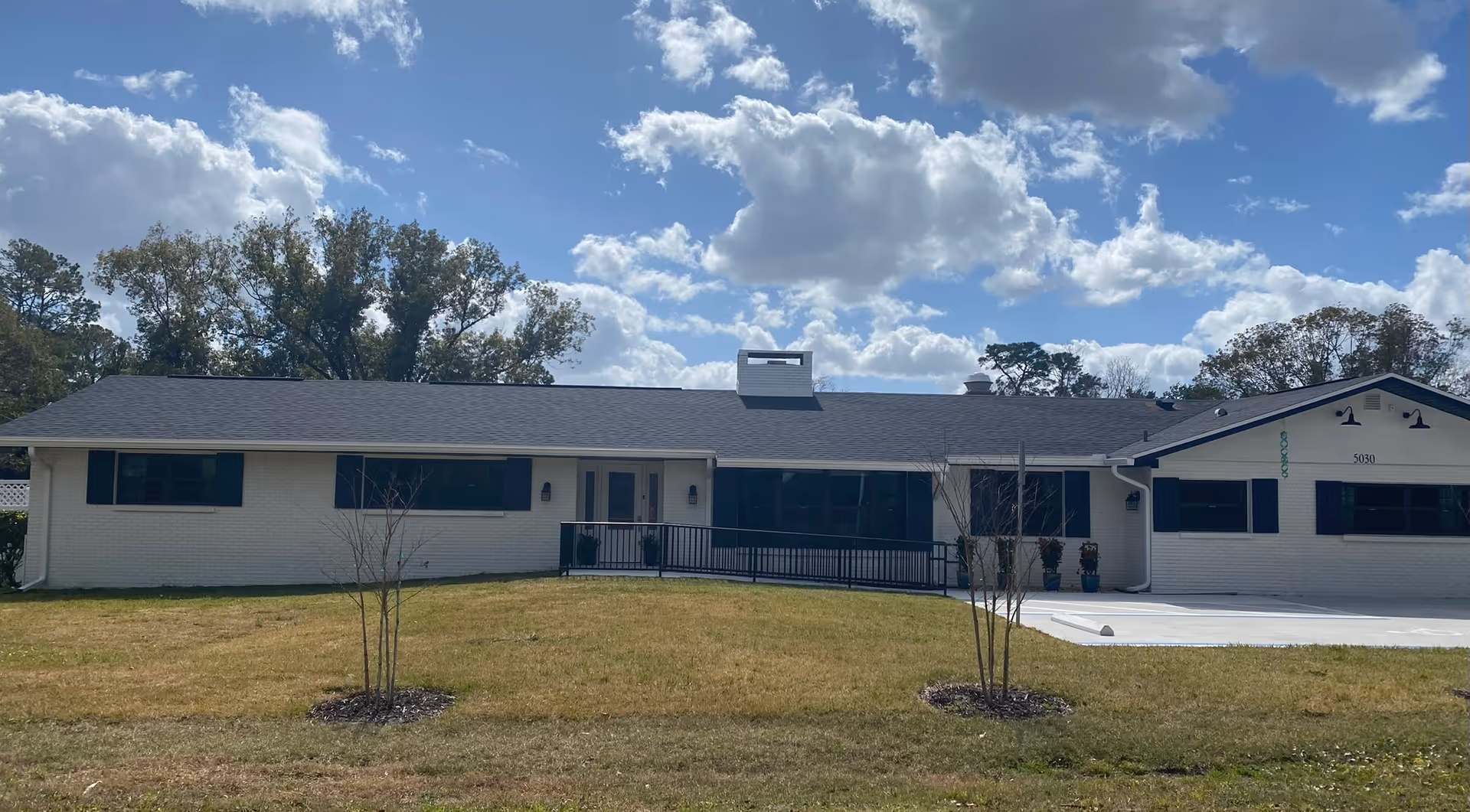 Single-story building with white brick exterior and black shutters under a partly cloudy blue sky. There is a ramp leading to the entrance and a small lawn with two young trees in front.