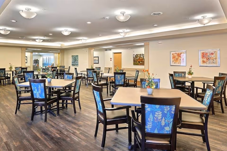 A spacious dining room with multiple square tables and chairs featuring blue floral upholstery. Each table has a small flower arrangement. The room has wood flooring, cream-colored walls, ceiling lights, and framed artwork on the walls.