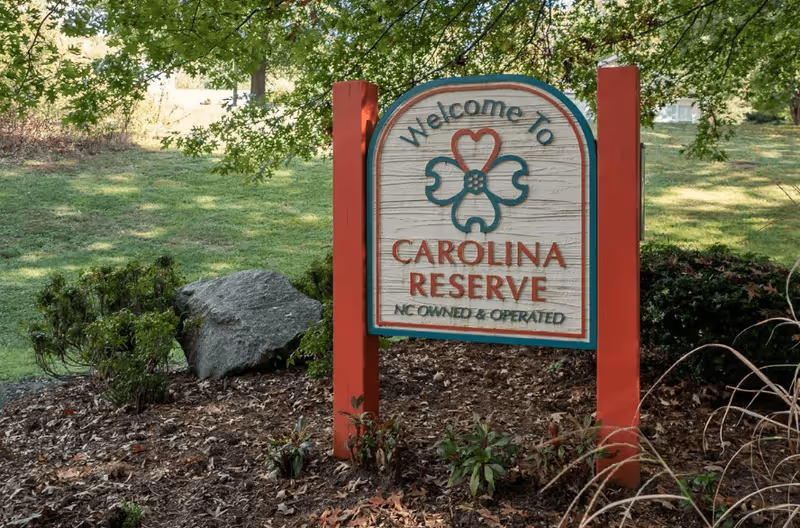 A wooden sign with red posts in a landscaped garden area that reads 'Welcome to Carolina Reserve NC Owned & Operated' with a decorative flower and heart design above the text. The background shows green grass, bushes, and trees.