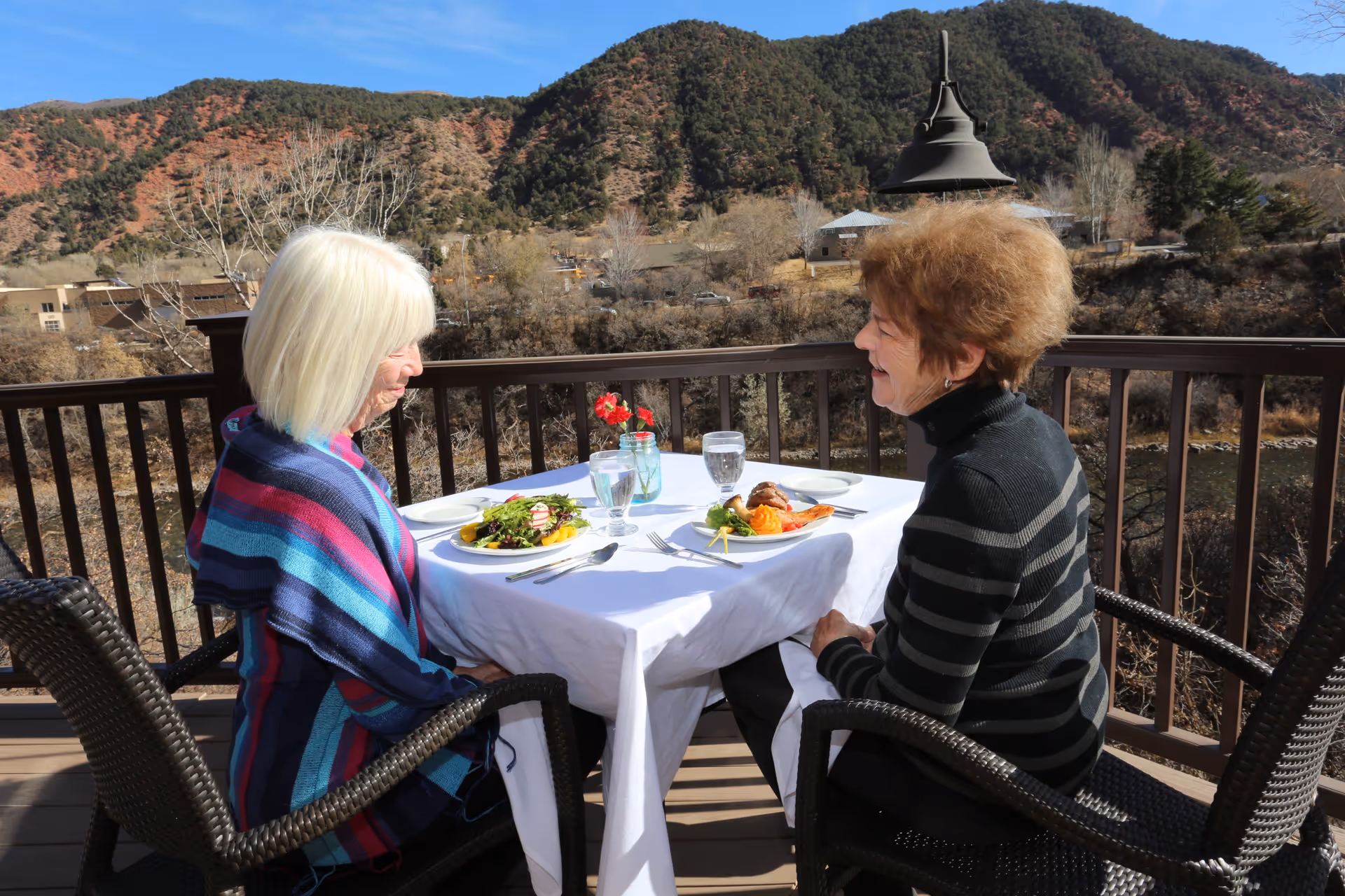 Two elderly women sitting at a table with a white tablecloth on an outdoor balcony, enjoying a meal with plates of food and glasses of water. Behind them is a scenic view of hills, trees, and a clear blue sky.