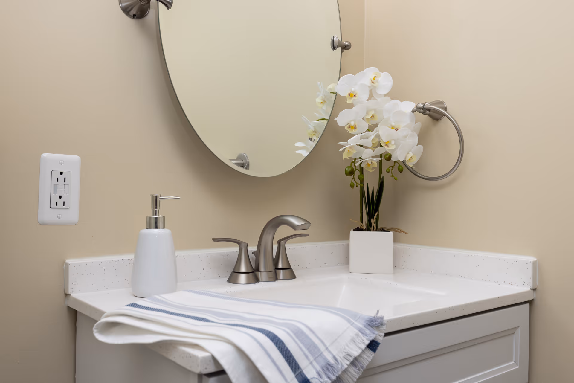 Bathroom vanity with a round mirror, faucet, soap dispenser, folded striped towel, and a potted white orchid.