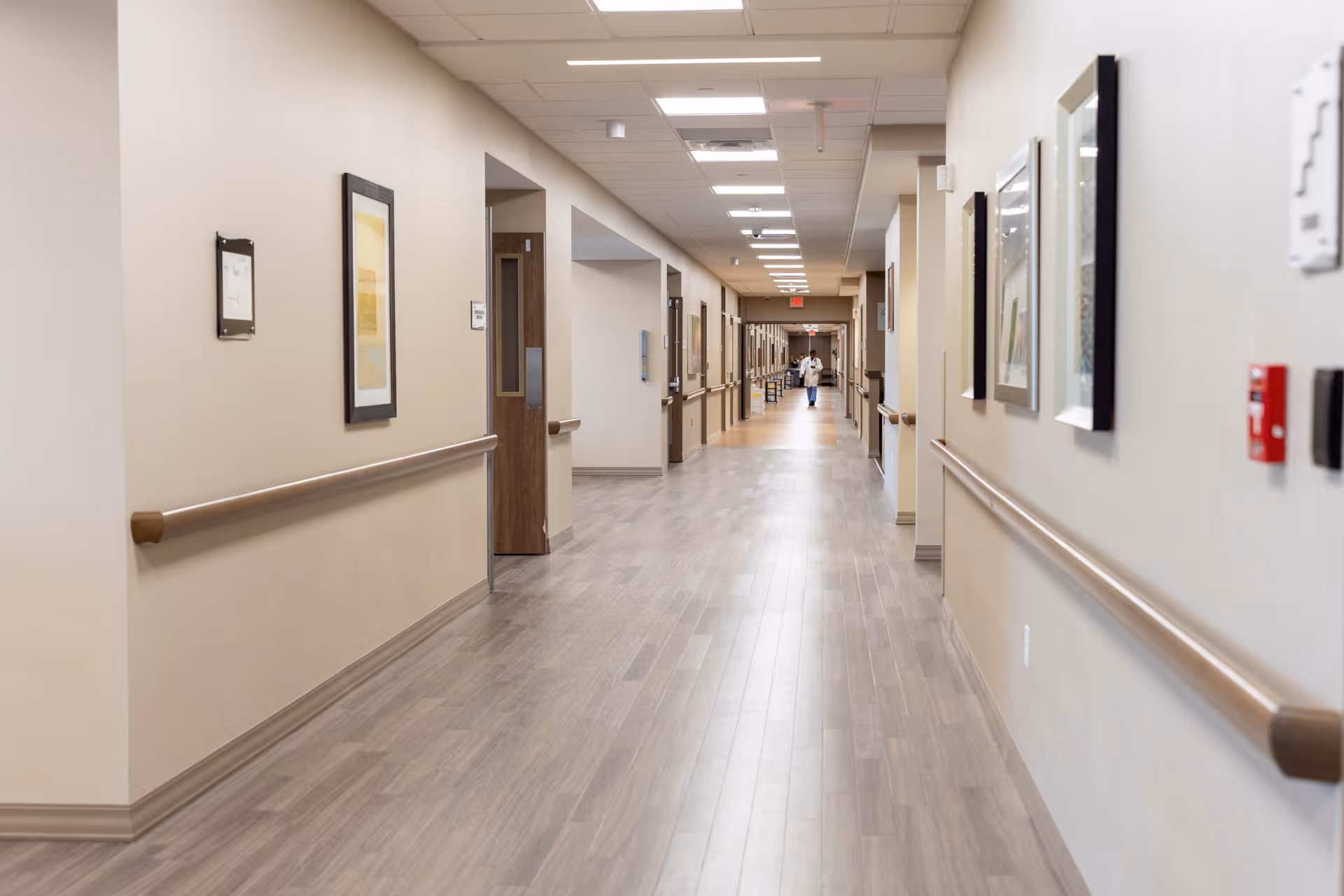 A long, clean hallway in a healthcare or senior living facility with light-colored walls, wooden handrails on both sides, framed artwork on the walls, and a person in a white coat walking in the distance.