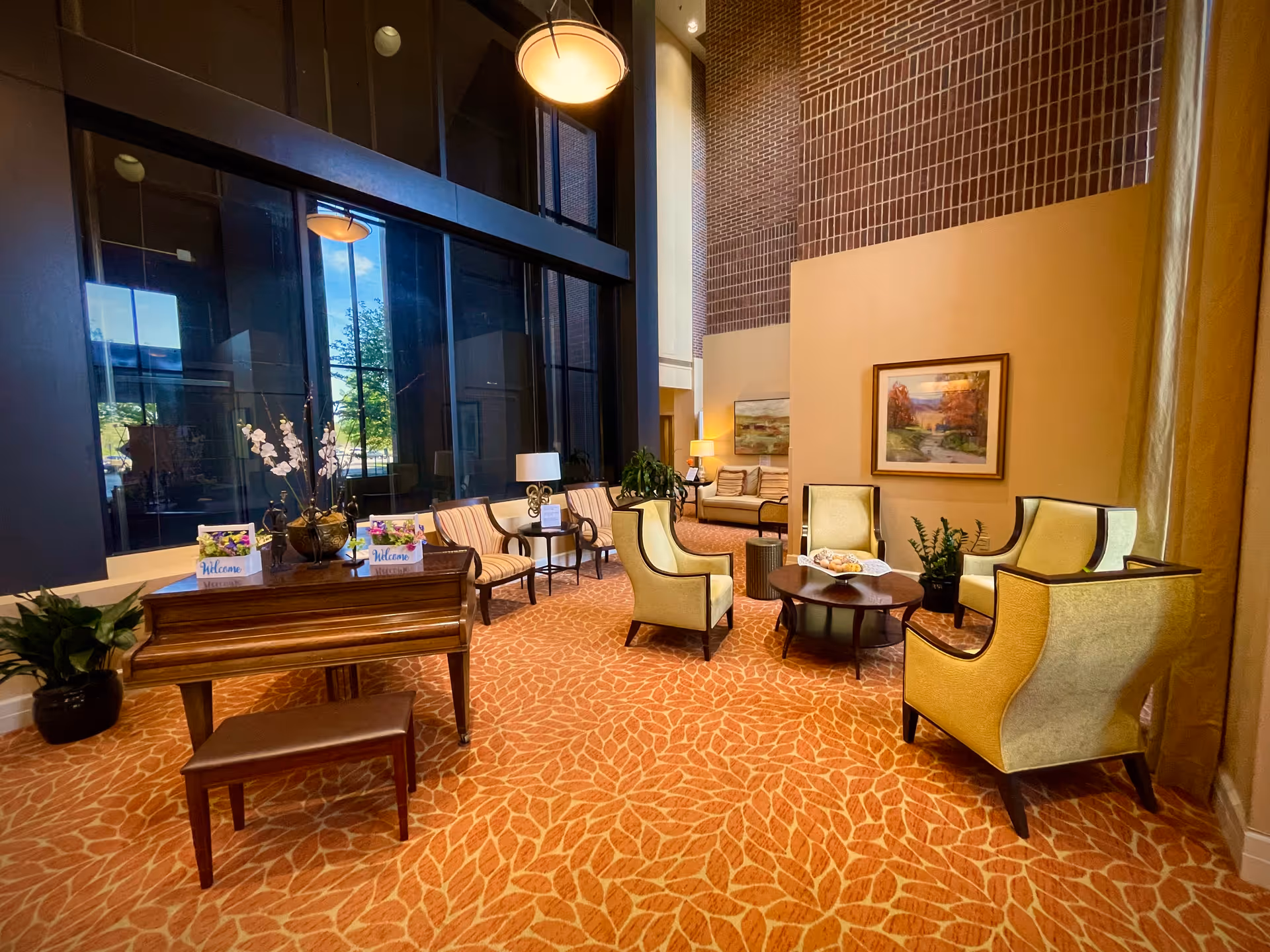 A spacious and well-lit living room area with large floor-to-ceiling windows, a wooden piano with a bench, several upholstered chairs arranged around a circular coffee table, decorative plants, and framed artwork on the walls. The carpet has a leaf pattern in warm tones, and there are table lamps providing additional lighting.