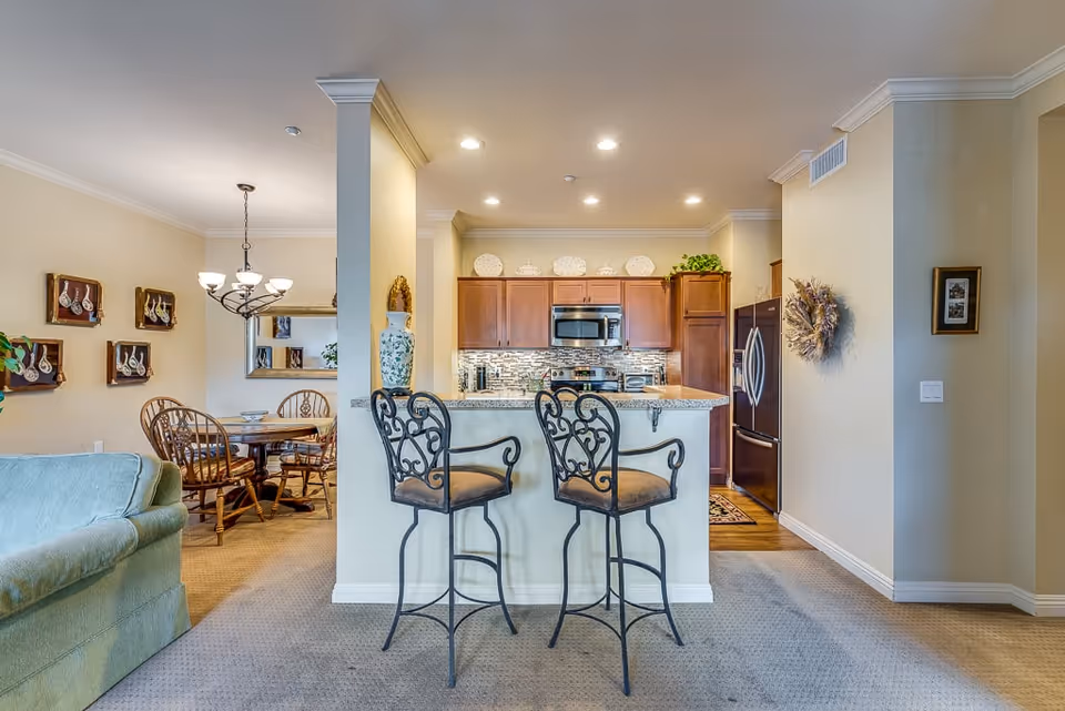 Interior view of a senior living facility showing a kitchen with wooden cabinets, stainless steel appliances, and a granite countertop with two ornate bar stools. To the left, there is a dining area with a round wooden table and chairs, a chandelier overhead, and decorative wall art. A green sofa is partially visible in the foreground.