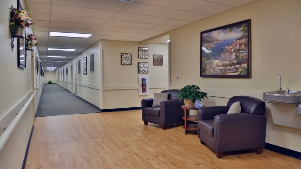 A long, well-lit hallway in a senior living facility with beige walls and a wooden floor area near the foreground. Two dark brown armchairs with cushions are placed around a small round wooden table holding a potted plant. A framed painting of a coastal scene hangs on the wall above the chairs. A water fountain is mounted on the wall to the right. The hallway extends into the distance with handrails and framed pictures on the walls.