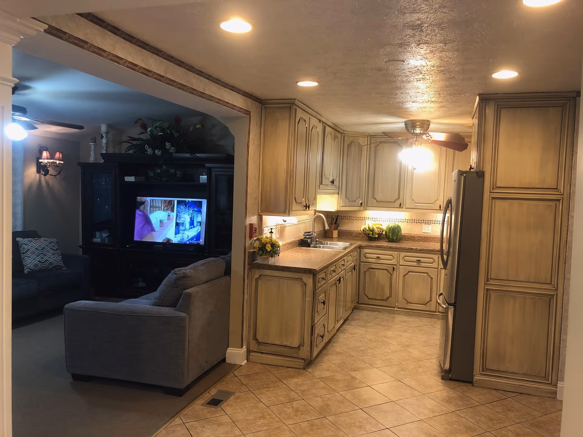Interior view showing a kitchen with wooden cabinets, a ceiling fan with lights, a stainless steel refrigerator, and a countertop with a watermelon and a fruit basket. Adjacent to the kitchen is a living room area with a gray sofa, a dark wooden entertainment center, and a TV turned on. The living room also has a ceiling fan and wall sconces with lampshades.