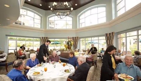 Seniors seated at round tables being served meals in a bright dining room with tall arched windows and a chandelier.