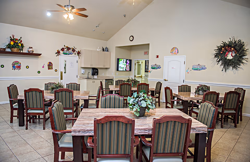 A dining room with multiple wooden tables and striped upholstered chairs arranged neatly. Each table has a small plant centerpiece. The room has beige walls decorated with wreaths, floral arrangements, and colorful wall hangings. There is a ceiling fan with lights, a wall-mounted TV, and a small kitchenette area with cabinets, a sink, and a paper towel dispenser. An exit sign is visible above a doorway.