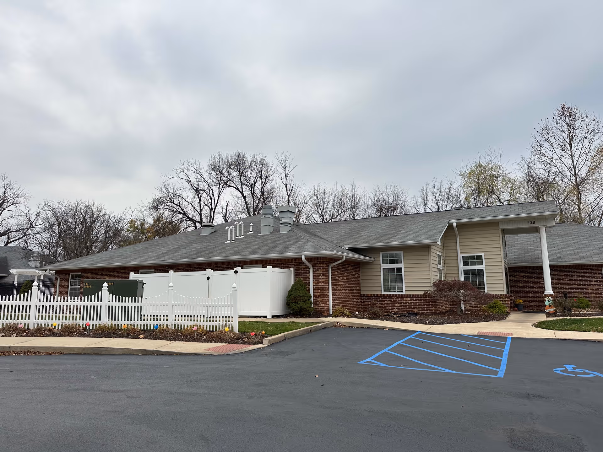 Exterior view of a single-story senior living facility building with brick and beige siding, white picket fence, and a handicapped parking space in front. The sky is overcast and there are leafless trees in the background.