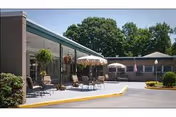 Outdoor patio area at a senior living facility with chairs, tables, and umbrellas. The building has a covered walkway with hanging plants and is surrounded by trees and greenery under a clear sky.