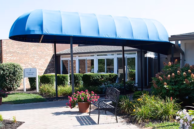 Outdoor entrance area of a facility with a blue canopy over a paved walkway, surrounded by green bushes, flowering plants, and a metal bench. A sign near the entrance reads 'Reception Center'.