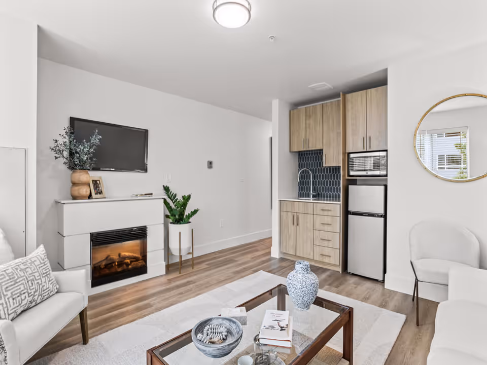 A modern living room with light-colored walls and wood flooring. The room features a white armchair with a patterned pillow, a white fireplace with a flat-screen TV mounted above it, and two potted plants. There is a small kitchenette with wooden cabinets, a sink, a microwave, and a mini refrigerator. A round mirror hangs on the wall next to a light gray chair. A glass coffee table with decorative items and books sits on a light area rug in the center of the room.