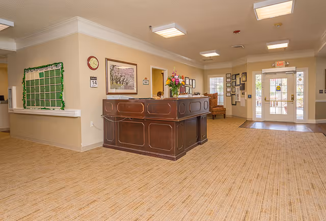 Well-lit senior living facility lobby with a wooden reception desk, seating area, bulletin board, and glass entrance doors.