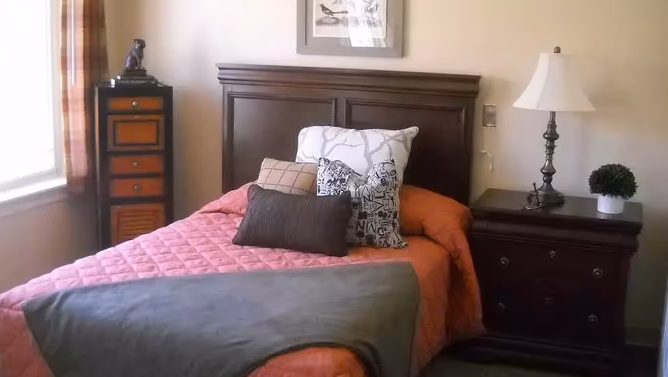 Cozy bedroom with a single bed topped with multiple pillows, a wooden headboard, a nightstand with a lamp, and a small dresser by the window.