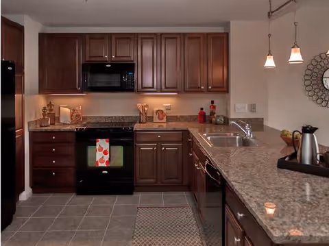 A modern kitchen with dark wooden cabinets, granite countertops, a black stove with a colorful towel hanging on the handle, a microwave above the stove, a double sink, and pendant lights hanging above the counter. The floor is tiled and there is a small rug in front of the stove.