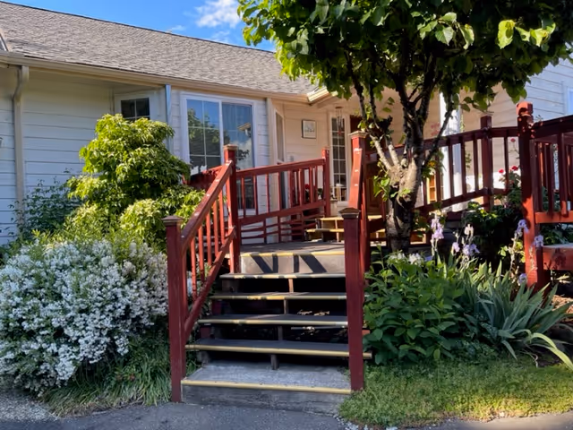 Front entrance of a house with red wooden steps and an accessibility ramp, bordered by green shrubs and flowering plants.