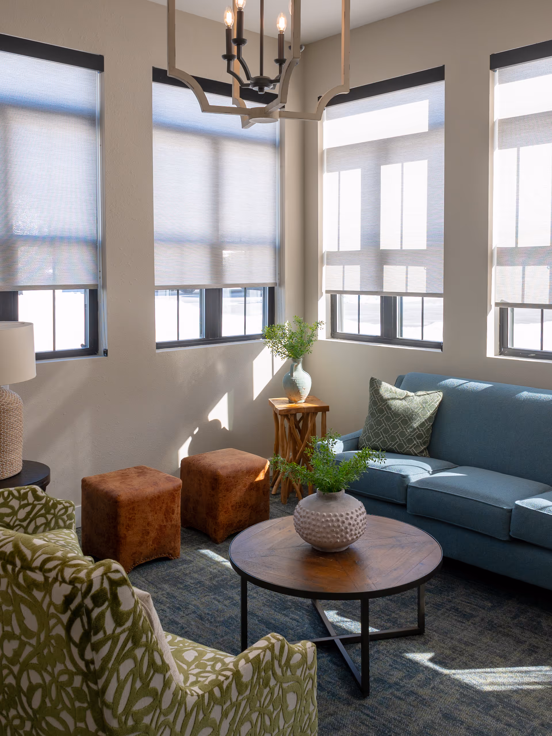 A bright living room with large windows covered by light-colored blinds. The room features a blue sofa with a patterned green pillow, two brown ottomans, a green patterned armchair, a round wooden coffee table with a decorative vase containing greenery, and a small wooden side table with another vase of greenery. A modern chandelier hangs from the ceiling.
