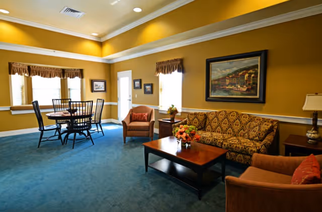 A cozy living room area with mustard yellow walls and blue carpet. The room features a patterned sofa, two armchairs with red cushions, a wooden coffee table with a floral arrangement, and a round dining table with four chairs near the windows. There are framed pictures on the walls and a table lamp on a side table.