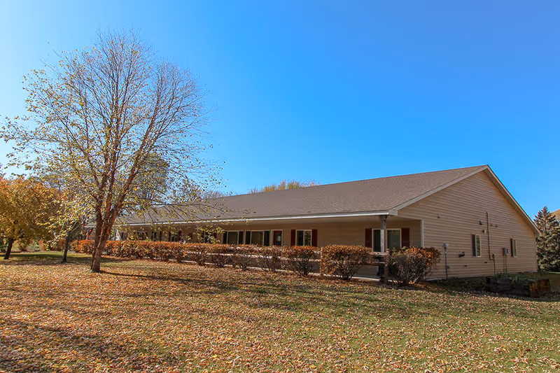 Single-story beige assisted living building with a covered porch, shrubs and trees on a grassy lot under a clear blue sky.